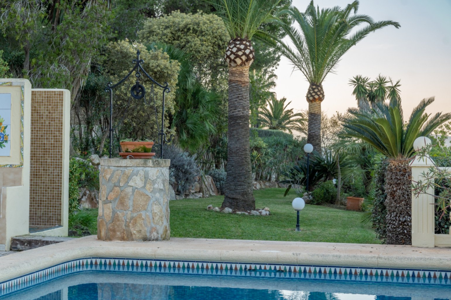 Jardín mediterráneo con césped, palmeras y pozo decorativo de piedra junto a la piscina. Incluye zona de ducha exterior alicatada y luminarias esféricas.