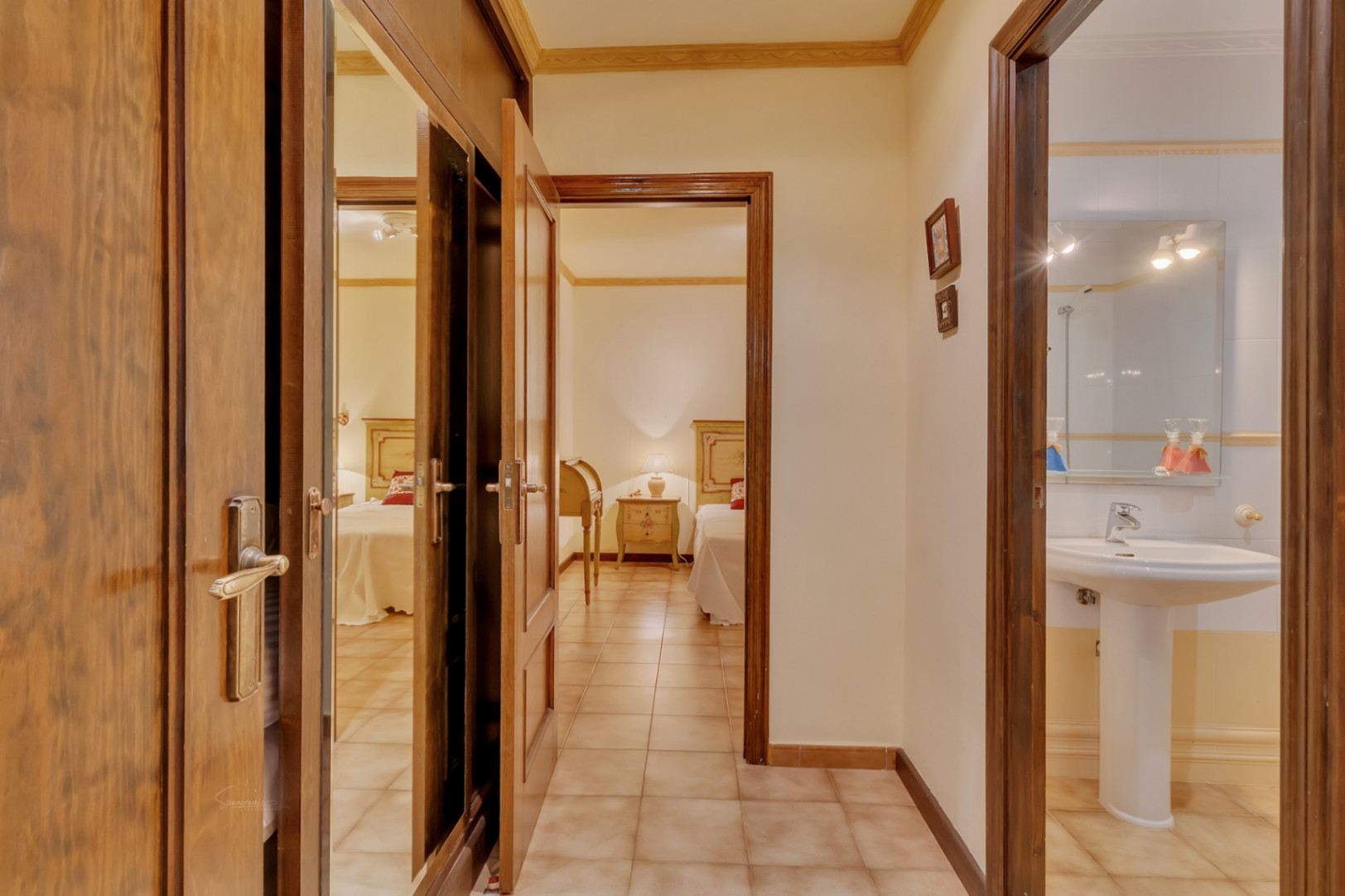 Pasillo distribuidor con armarios empotrados de madera y espejos, que conecta un dormitorio con camas individuales y un baño con lavabo de pedestal.