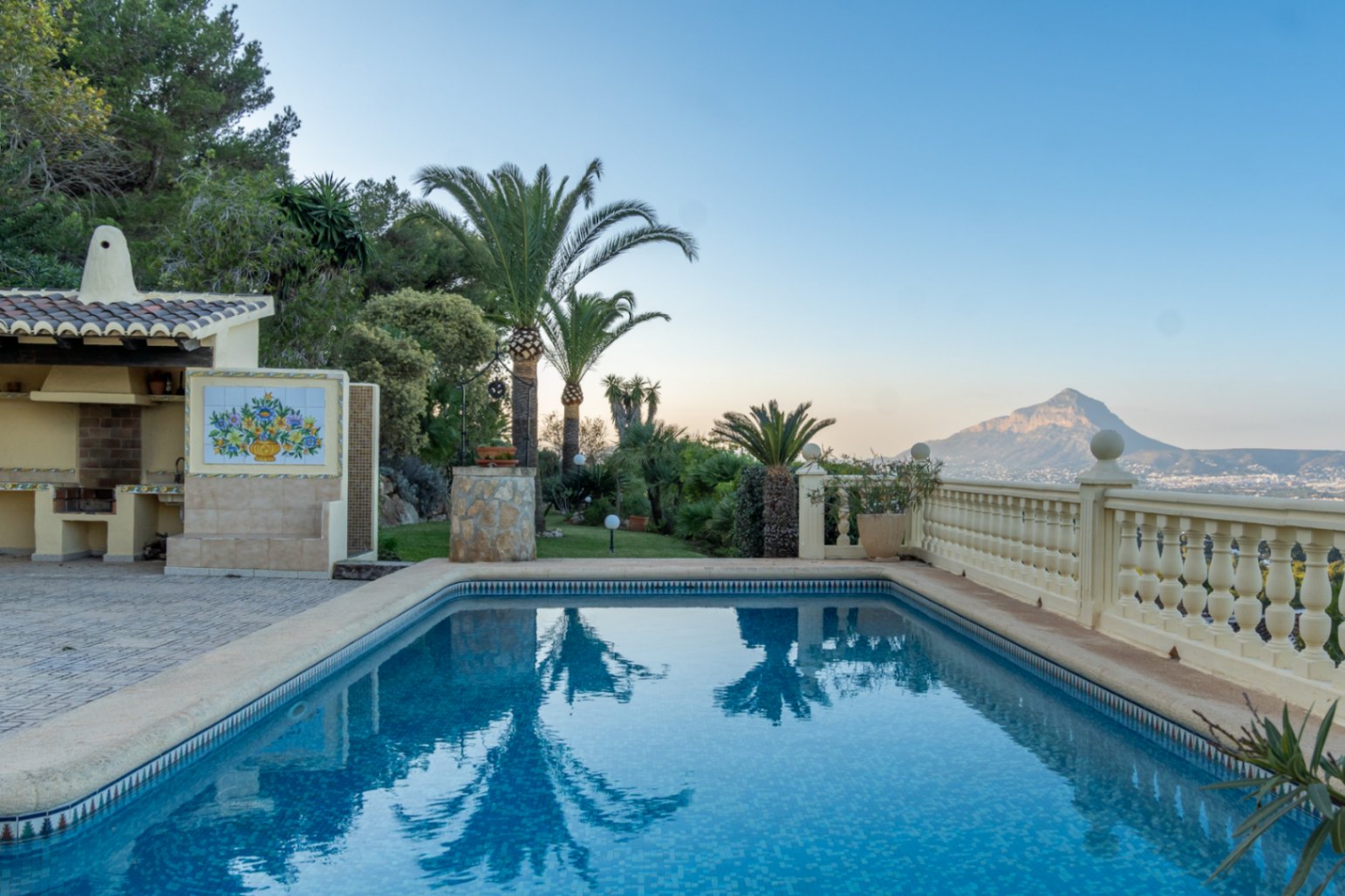 Piscina rectangular con azulejos azules, zona de barbacoa techada y vistas panorámicas al Montgó y la bahía desde la terraza con balaustrada clásica.