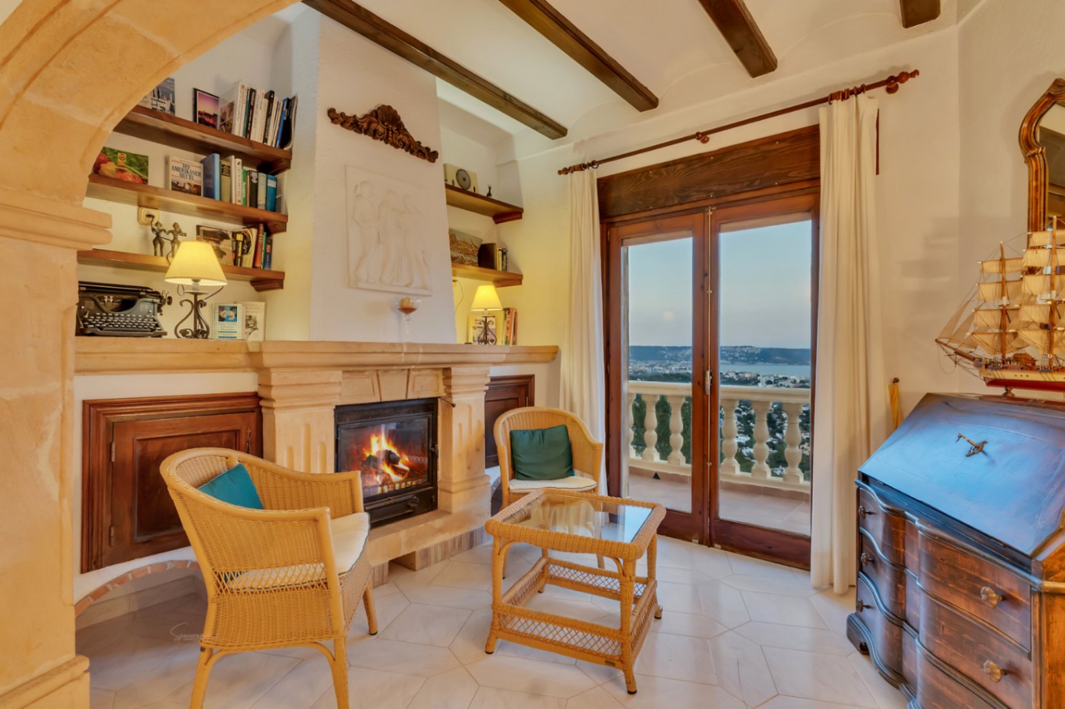 Salón con chimenea de piedra, vigas de madera y mobiliario de mimbre. Las puertas acristaladas conectan con la terraza y ofrecen vistas a la bahía de Jávea.