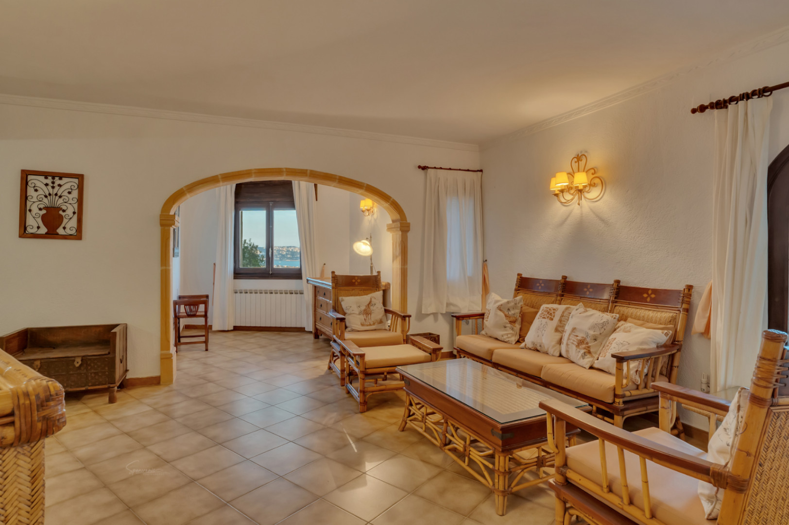 Salón con mobiliario de bambú y arco de piedra tosca que conecta con una estancia con vistas al mar. Suelos cerámicos y apliques de pared de estilo clásico.