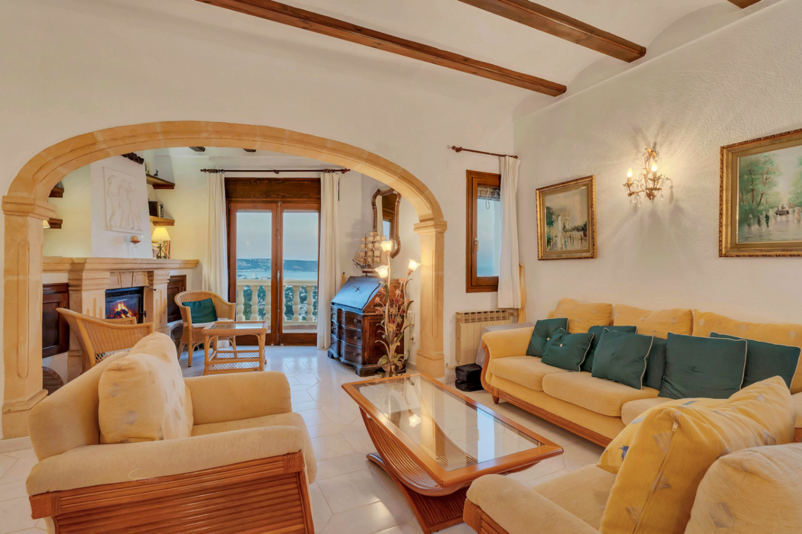Salón de estilo mediterráneo con vigas de madera, gran arco de piedra tosca y chimenea. Incluye acceso directo a la terraza con vistas panorámicas al mar.