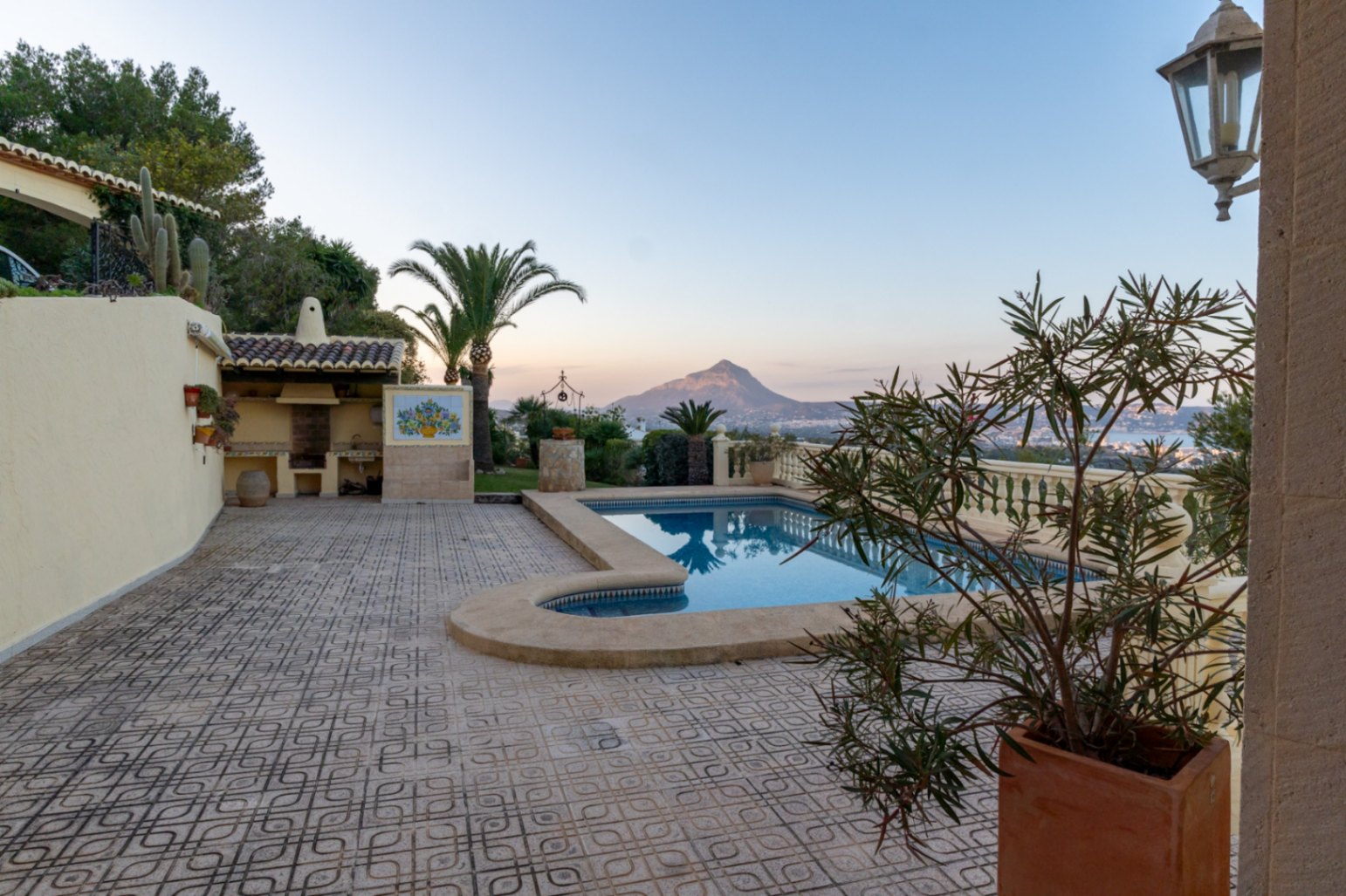 Terraza exterior con suelo cerámico decorado, piscina rectangular y zona de barbacoa cubierta. Ofrece vistas despejadas al Montgó y la bahía bajo una iluminación de atardecer.