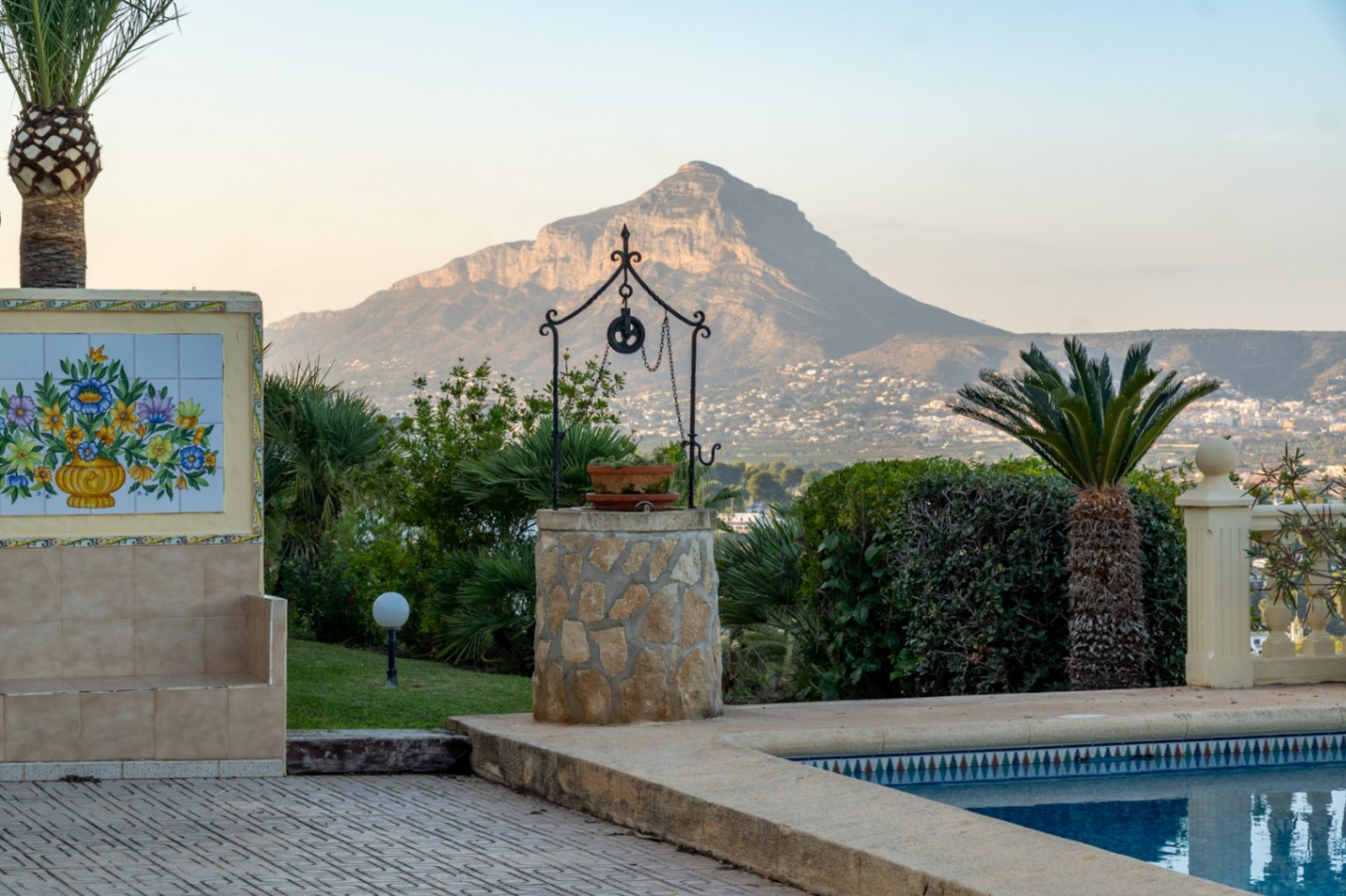 Terraza junto a la piscina con pozo decorativo de piedra y mural cerámico, ofreciendo vistas panorámicas al Montgó y la costa de Jávea.