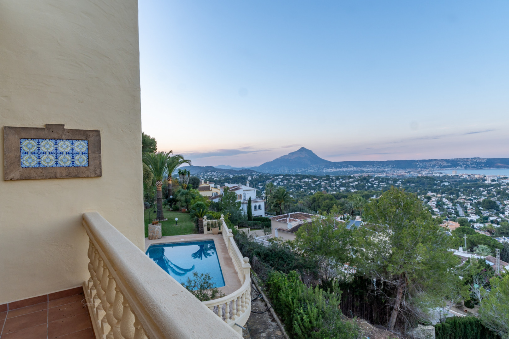 Terraza superior con balaustrada de piedra y azulejo decorativo, ofreciendo vistas a la piscina privada, el jardín mediterráneo y el perfil del Montgó sobre la bahía de Jávea.