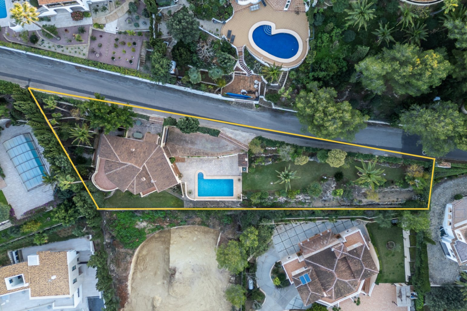 Vista aérea cenital de la propiedad delimitada, mostrando la distribución de la villa, la piscina rectangular, las zonas ajardinadas con palmeras y el acceso privado.