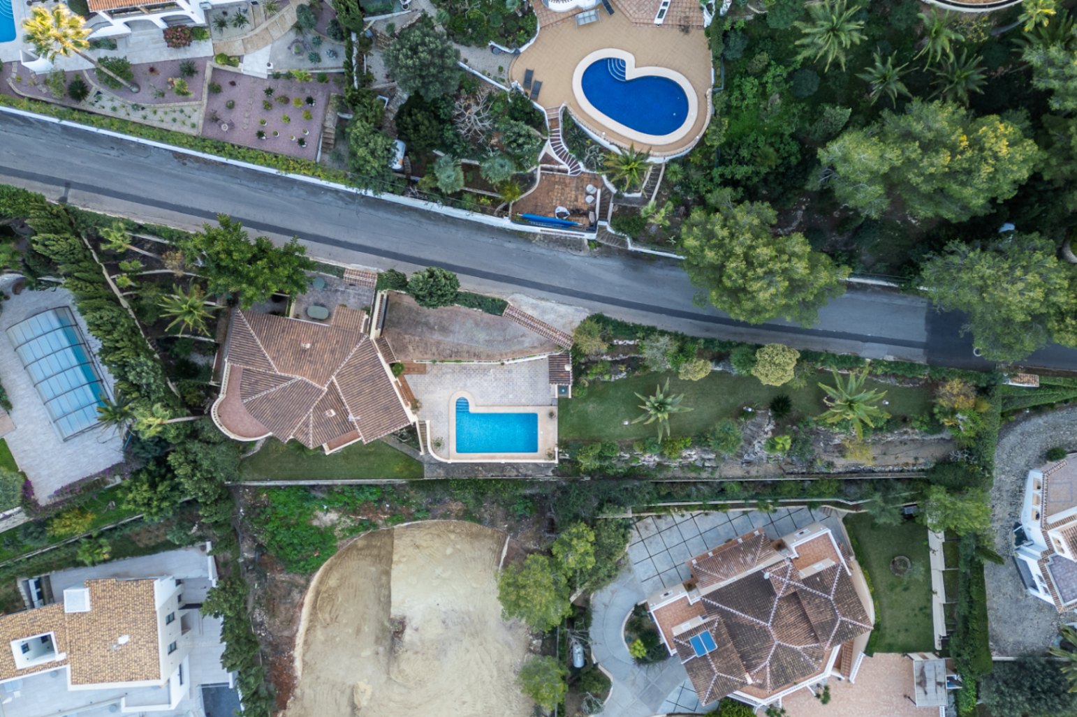 Vista aérea cenital de la villa que muestra la distribución de la parcela, la piscina rectangular, las terrazas pavimentadas y el jardín mediterráneo con palmeras.
