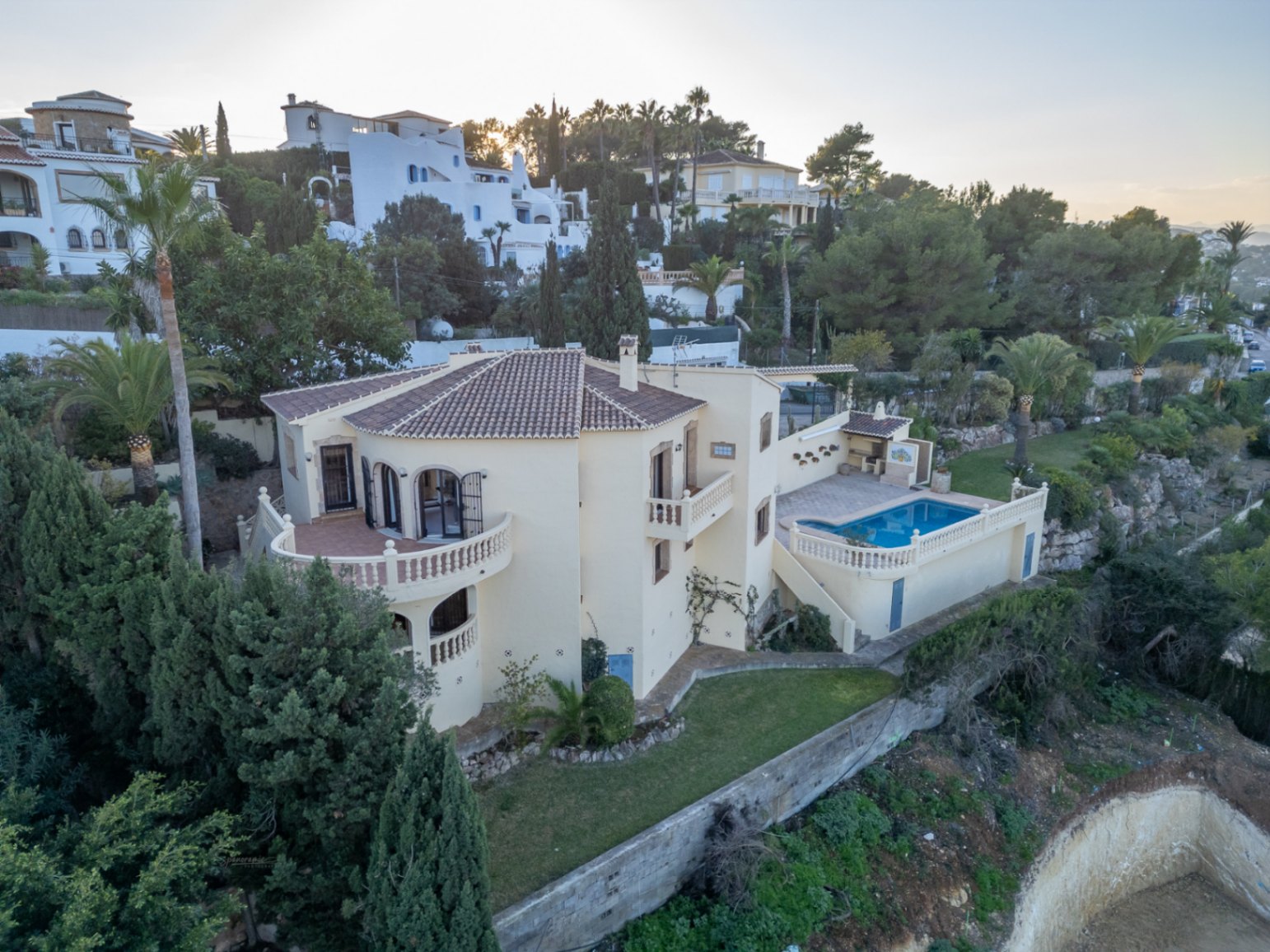 Vista aérea de la villa con fachada color crema, tejado de teja y amplias terrazas con balaustradas. Incluye zona de piscina, barbacoa y jardín mediterráneo.