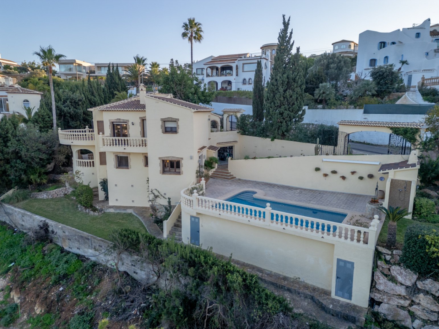 Vista exterior de la villa mediterránea con piscina privada, terrazas con balaustradas blancas y fachada en tonos crema con detalles en piedra natural.