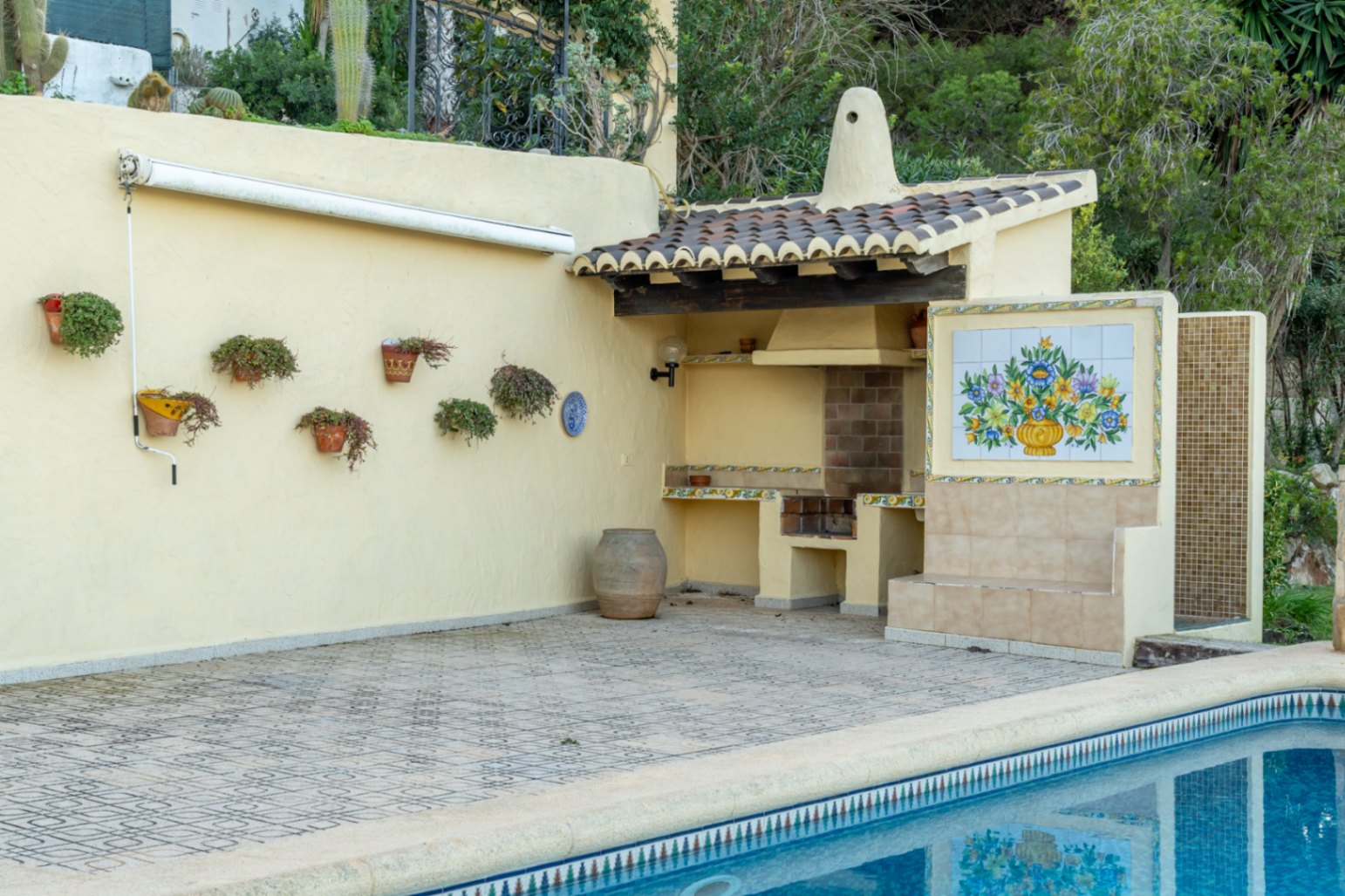 Zona de barbacoa exterior junto a la piscina, con tejadillo de teja cerámica, mural decorativo de azulejos y suelo de gres estampado en tonos neutros.