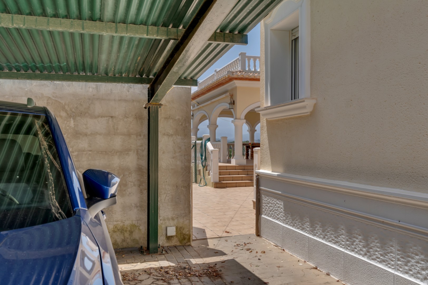 Área de estacionamiento cubierto con marquesina metálica junto a la entrada principal, con acceso directo a la terraza de arcos y la naya de la villa.