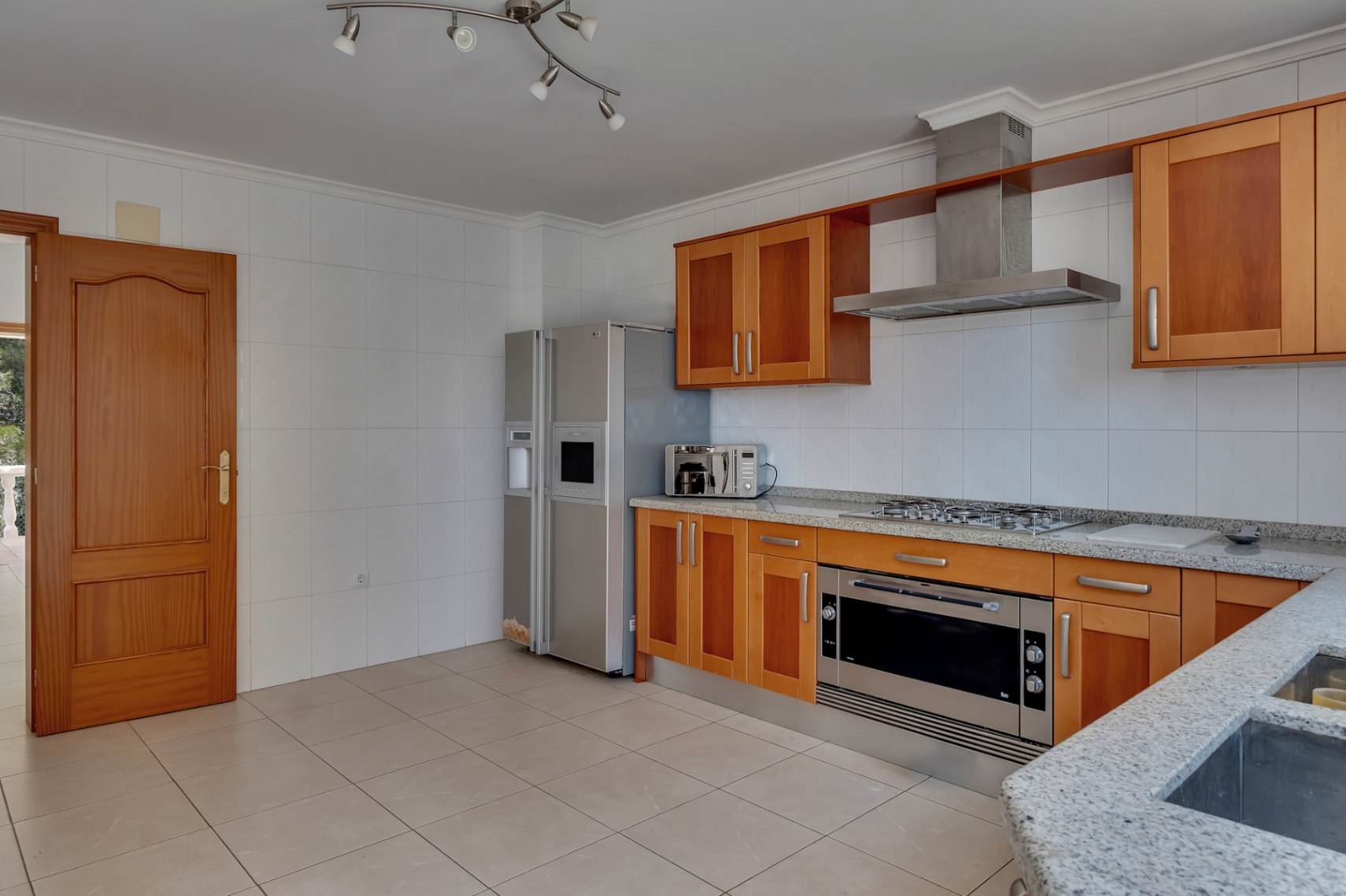 Cocina equipada con mobiliario de madera, encimeras de granito gris y electrodomésticos de acero inoxidable, incluyendo un frigorífico americano y horno de gran formato.