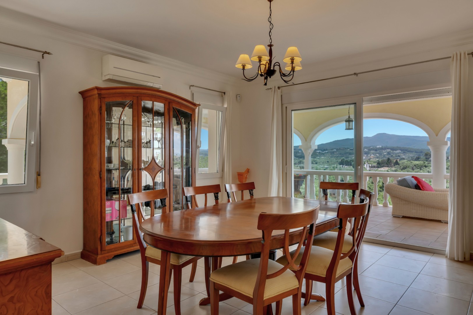Comedor con mesa ovalada de madera, vitrina clásica y acceso directo a la naya. Grandes ventanales ofrecen vistas panorámicas al valle y al Montgó.