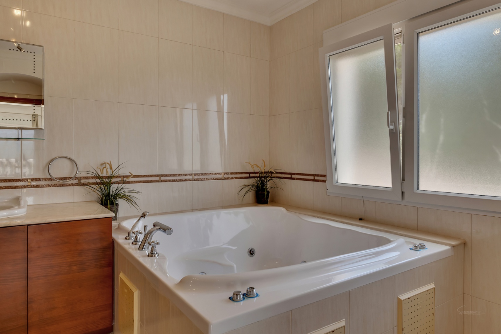 Cuarto de baño con bañera de hidromasaje integrada, revestimiento cerámico en tonos crema con cenefa decorativa y ventana de doble acristalamiento con vidrio traslúcido.