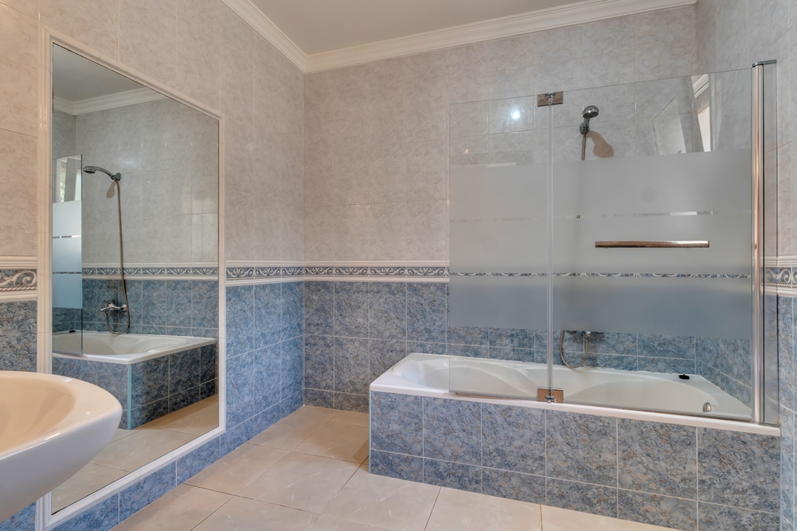 Cuarto de baño con bañera integrada, mampara de cristal parcial y revestimiento de azulejos en tonos azules con cenefa decorativa y un gran espejo.