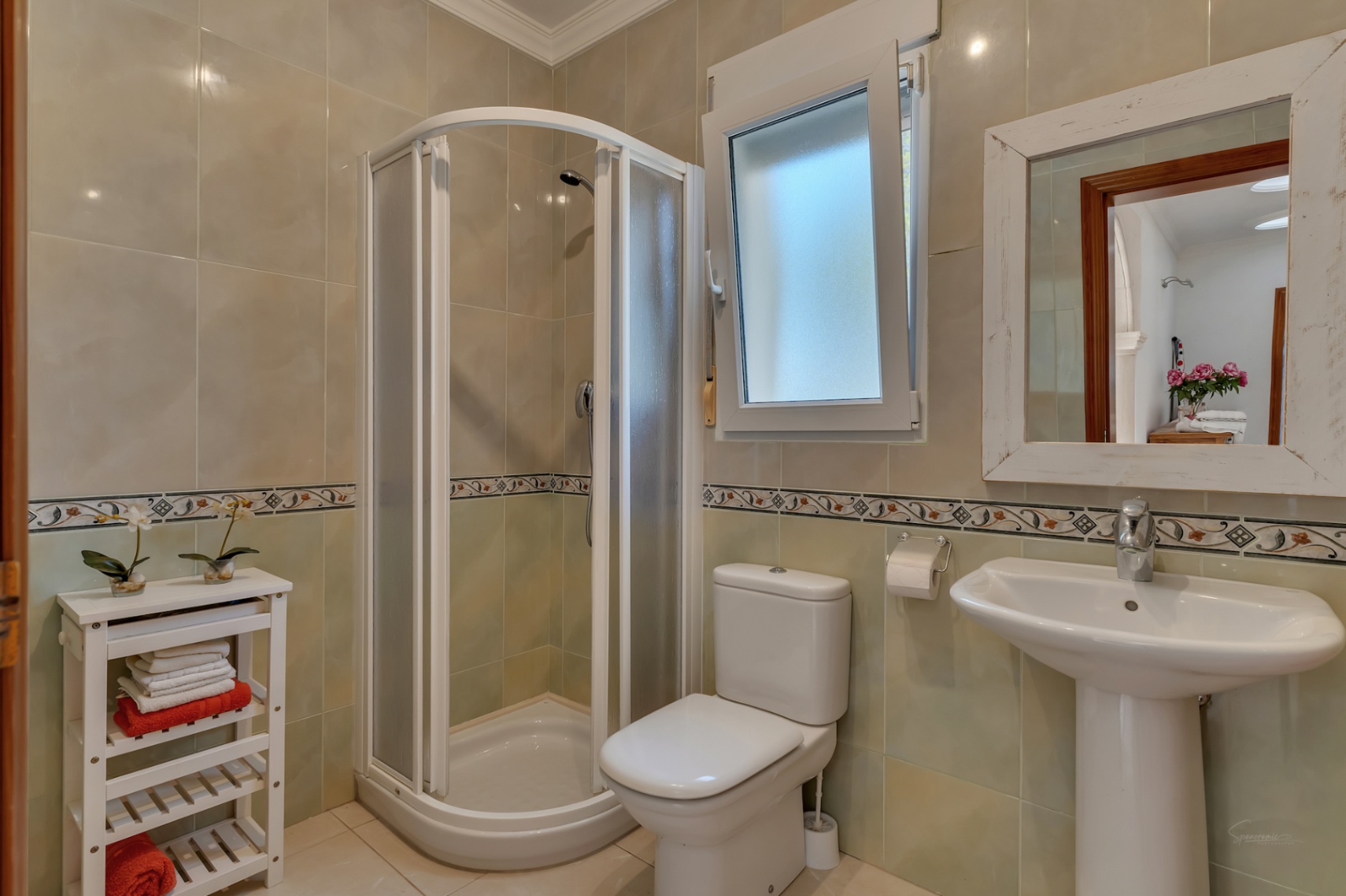 Cuarto de baño con ducha de esquina de puertas curvas, lavabo de pedestal blanco y paredes alicatadas con cenefa decorativa.