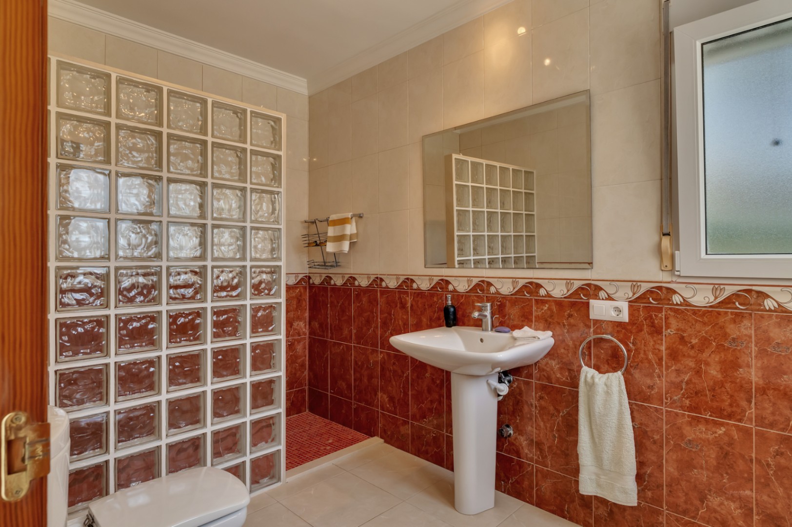 Cuarto de baño con lavabo de pedestal, espejo de pared y una ducha delimitada por bloques de vidrio. Destacan los azulejos de mármol rojizo y el suelo de mosaico en la zona de ducha.