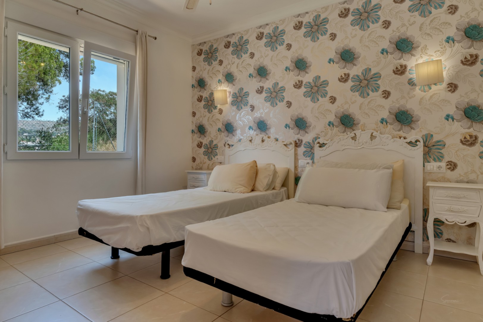 Dormitorio con dos camas individuales, cabeceros blancos de estilo clásico y papel pintado floral. Dispone de suelos cerámicos y ventana con vistas al entorno natural.