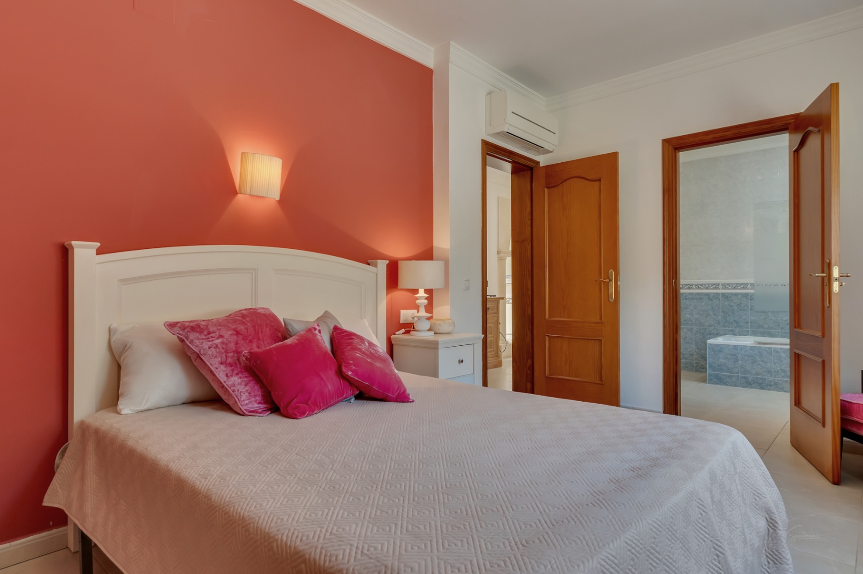 Dormitorio con pared de acento terracota, aire acondicionado split y acceso a un baño en suite con azulejos azules y bañera integrada.