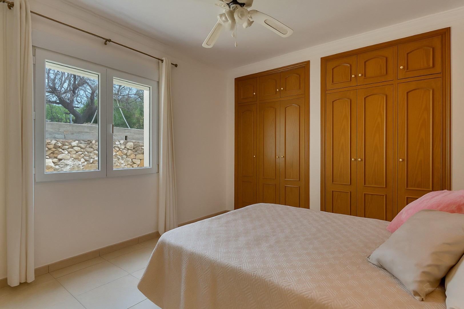 Dormitorio luminoso con armarios empotrados de madera, ventilador de techo y ventana con vistas al jardín y muro de piedra natural.