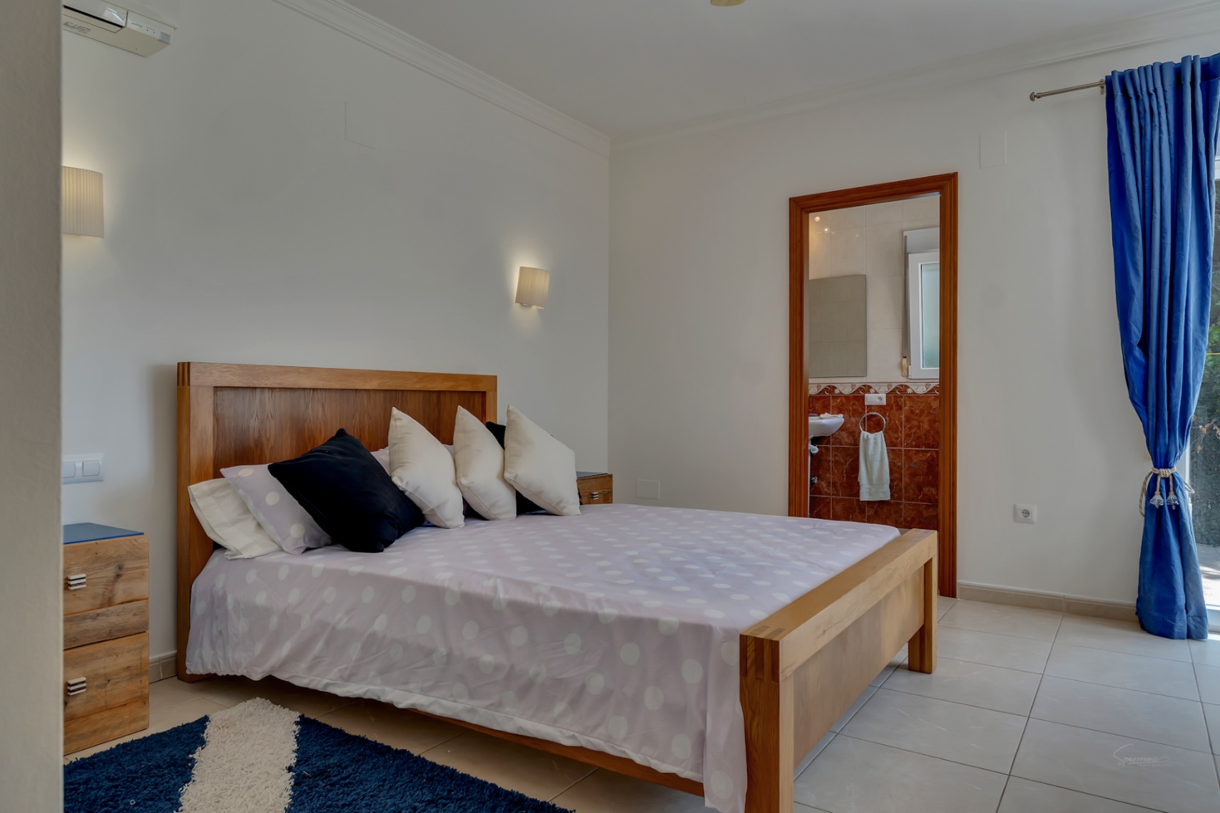 Dormitorio luminoso con cama de madera maciza, suelos de gres, aire acondicionado y acceso directo a un baño en suite con azulejos de mármol rojizo.