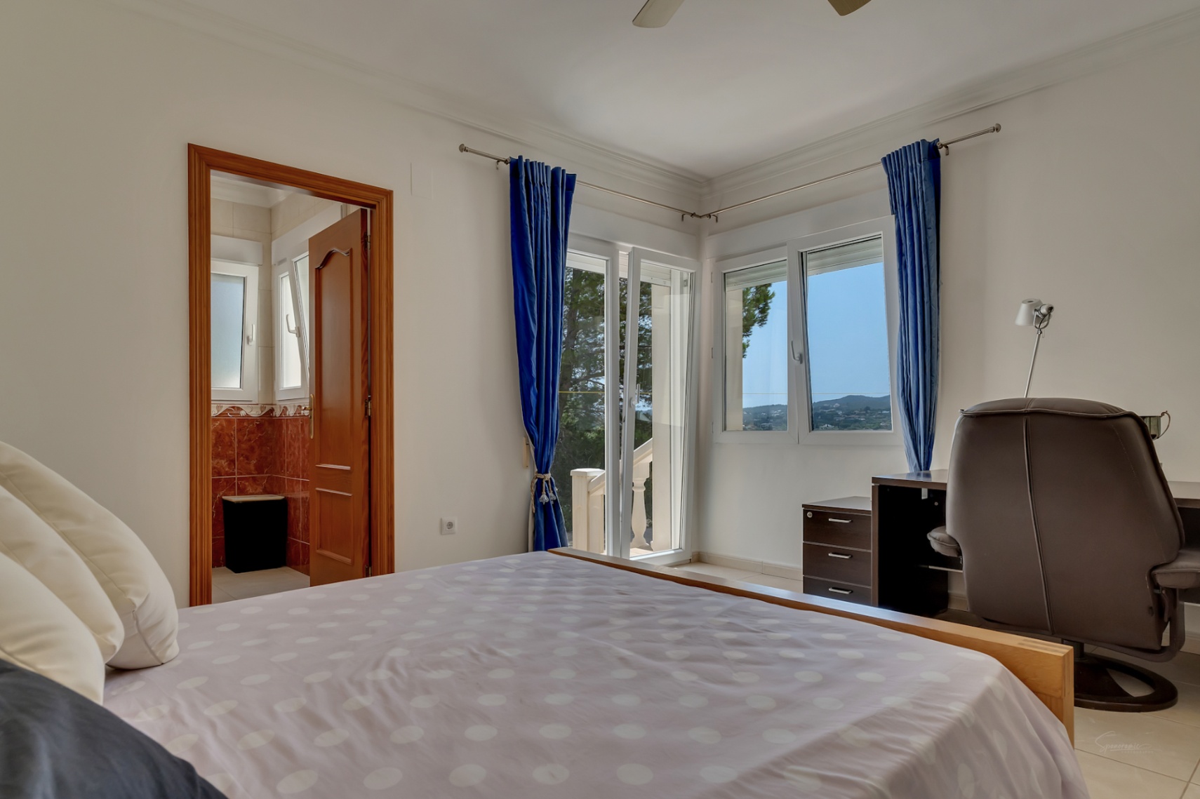 Dormitorio luminoso con zona de trabajo, acceso directo a la terraza y baño en suite. Destacan las vistas al valle a través de los ventanales con cortinas azules.