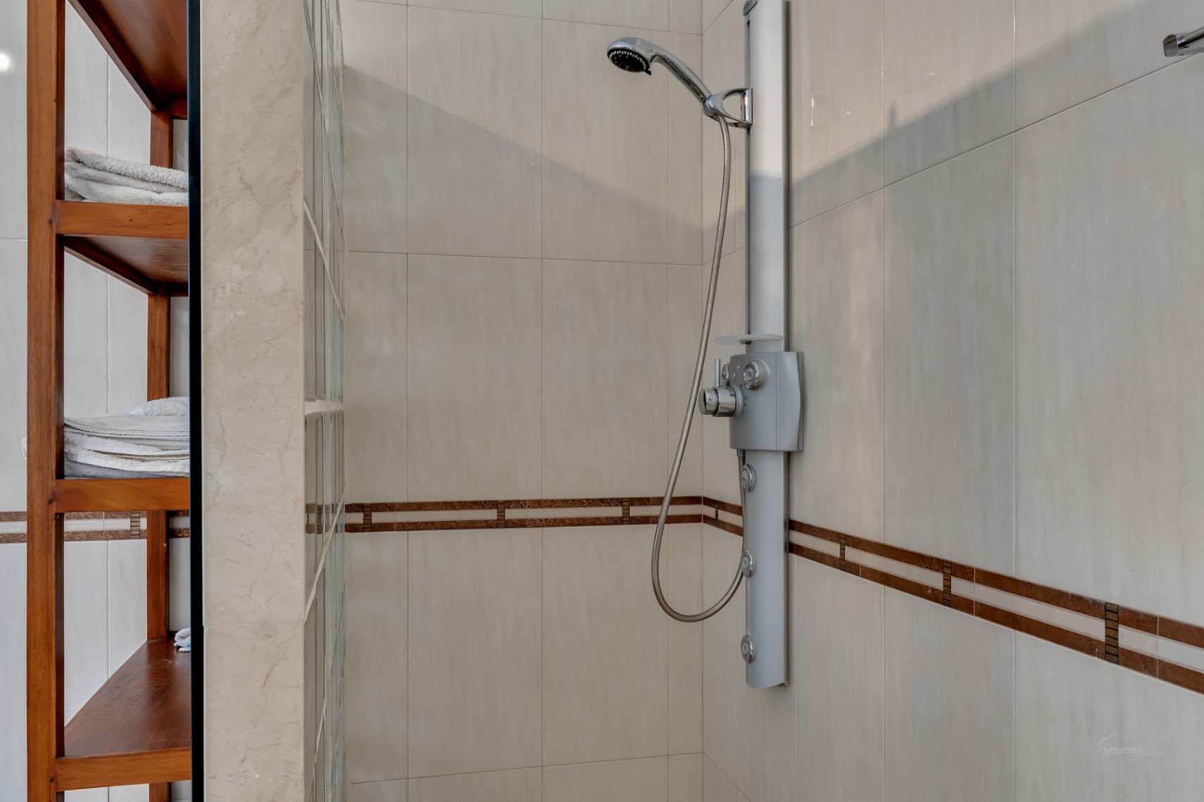 Ducha moderna con columna de hidromasaje, revestimiento cerámico en tonos crema con cenefa decorativa y estantería de madera integrada junto a pared de pavés.