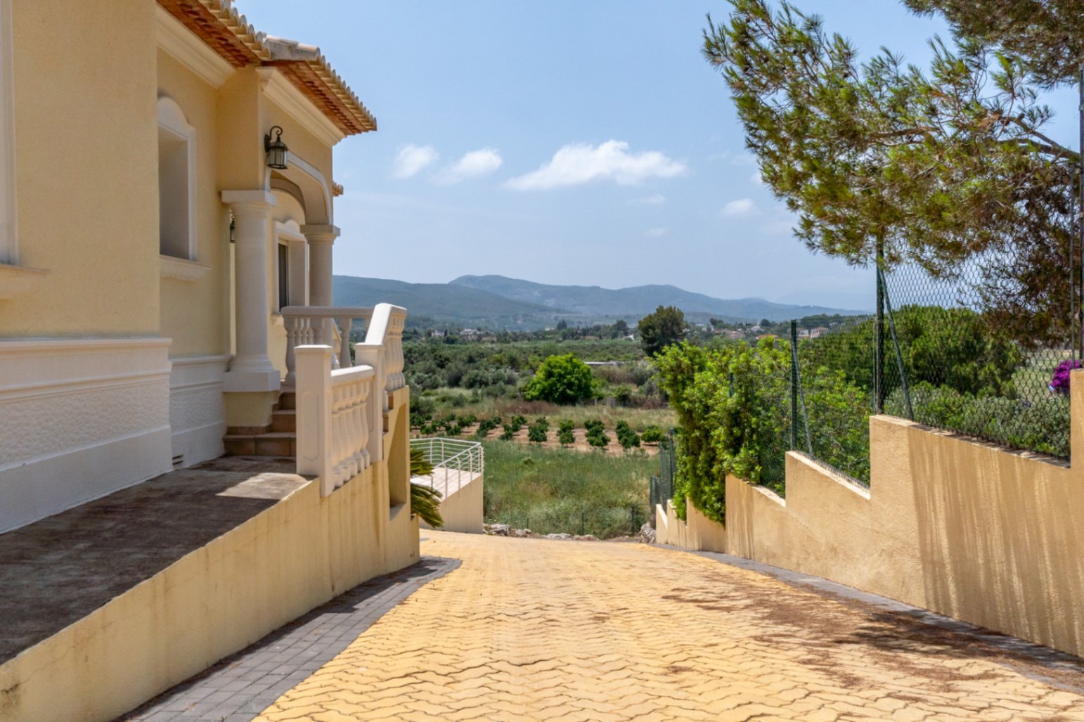Entrada de la villa con camino pavimentado descendente y porche con columnas, ofreciendo vistas despejadas al valle y las montañas circundantes.