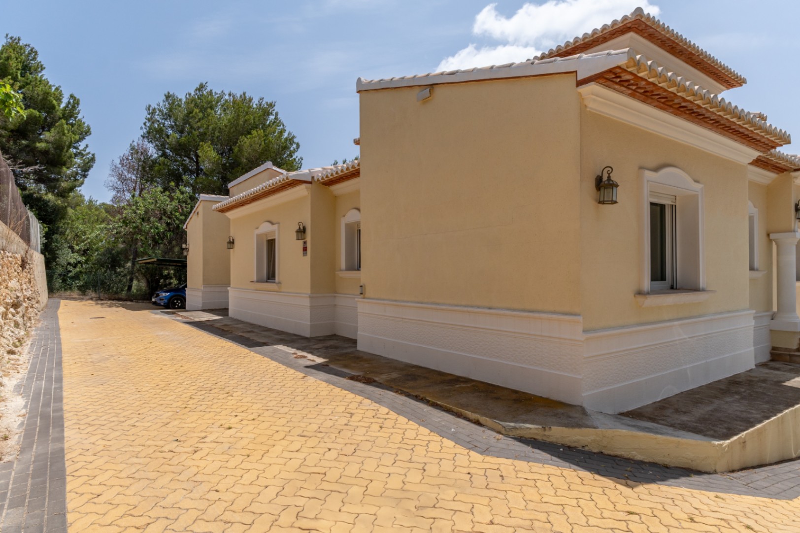 Fachada exterior en tonos ocres con molduras blancas, camino pavimentado y zona de aparcamiento cubierto rodeada de pinos mediterráneos.
