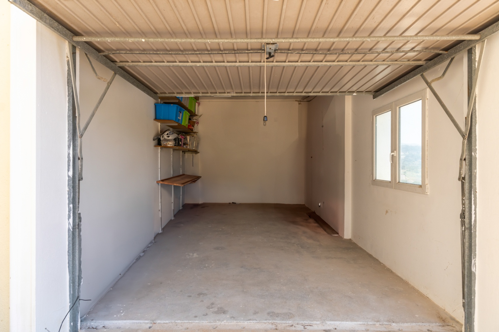 Garaje cerrado con suelo de hormigón, paredes blancas y ventana lateral. Incluye estanterías de almacenamiento y puerta metálica basculante.