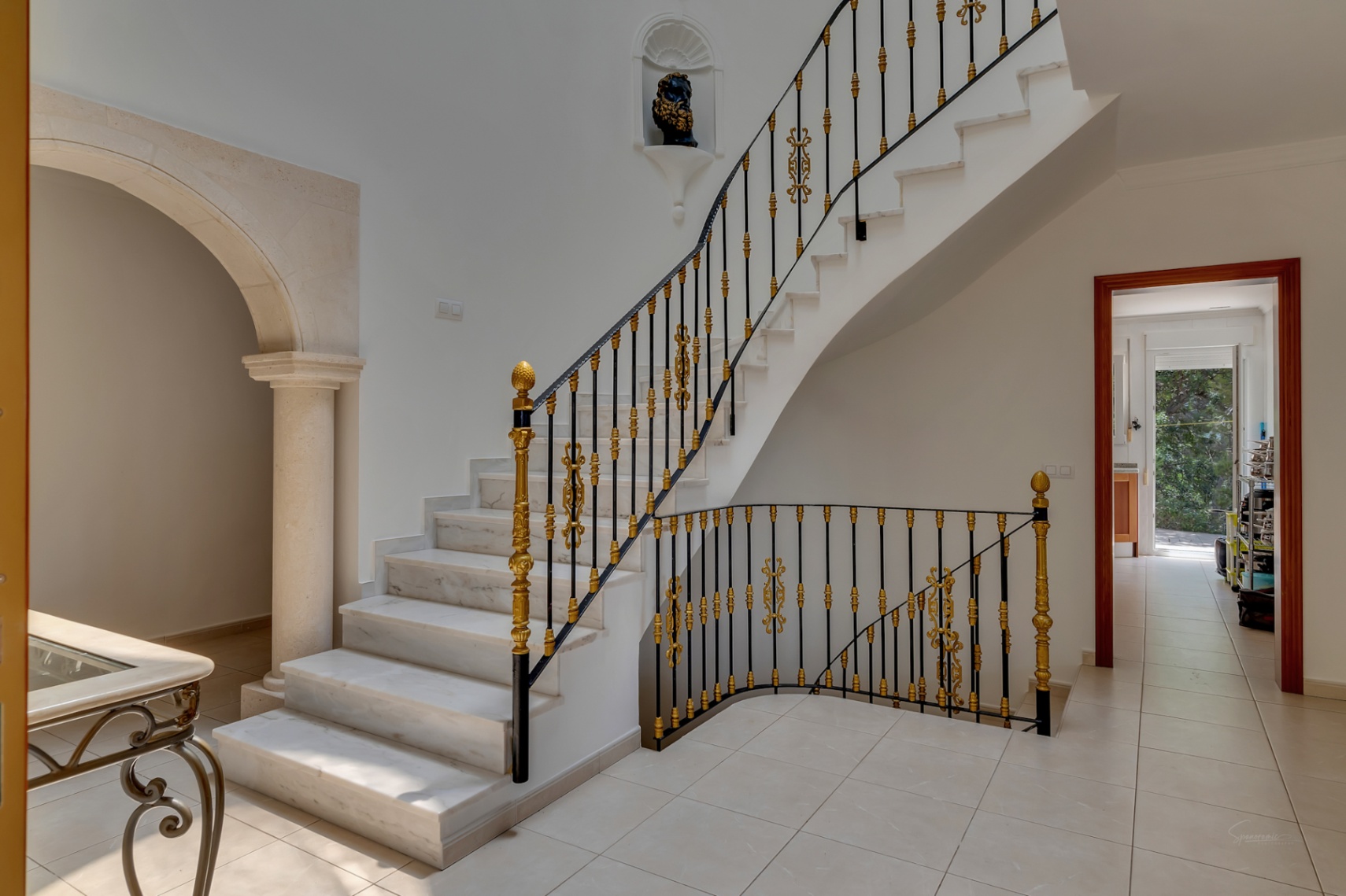 Hall de entrada con escalera de mármol, barandilla de hierro forjado con detalles dorados y un arco de piedra tallada con columna clásica.