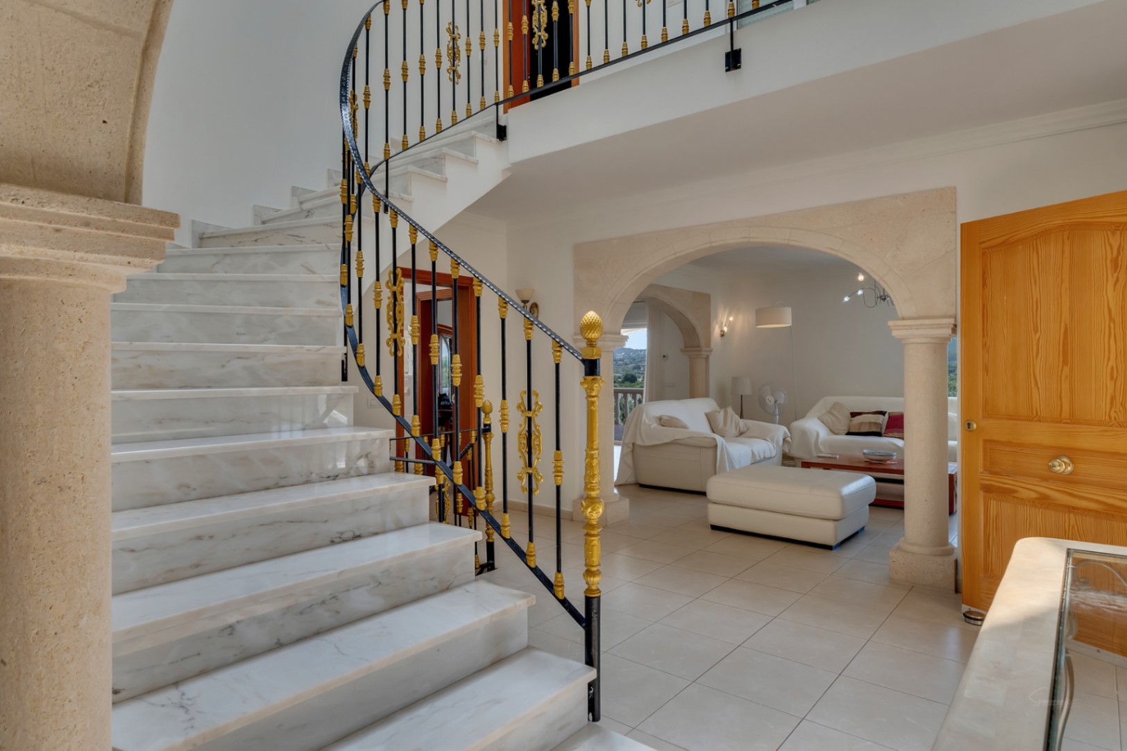 Hall de entrada con escalera de mármol y barandilla ornamental, conectado al salón mediante arcos de piedra natural y suelos cerámicos claros.