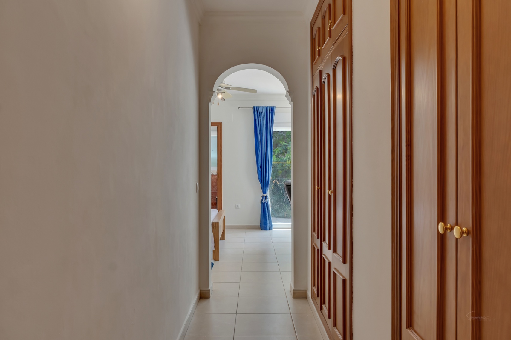Pasillo con amplios armarios empotrados de madera y suelo cerámico, que conecta mediante un arco con un dormitorio luminoso con ventilador de techo y cortinas azules.