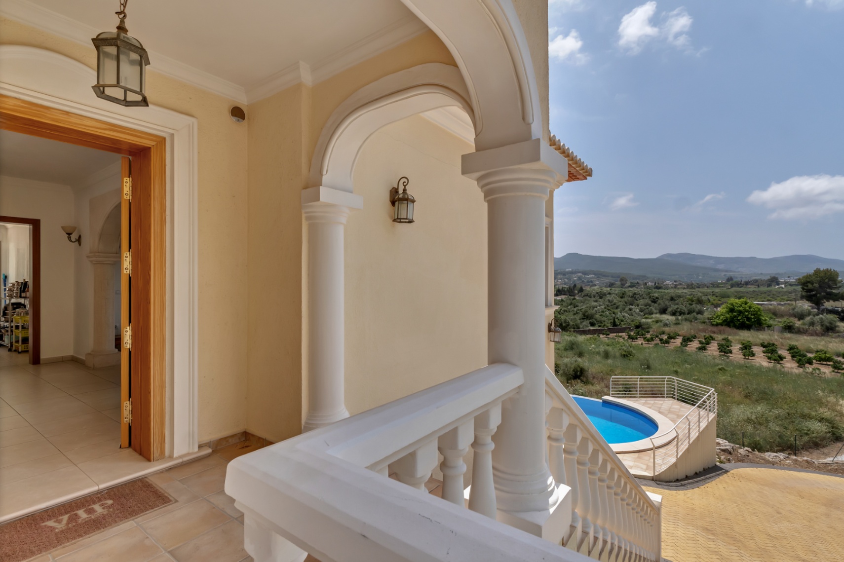 Porche de entrada con arcos y columnas blancas que ofrece vistas panorámicas al valle y la piscina circular. Suelo de gres y puerta principal de madera.