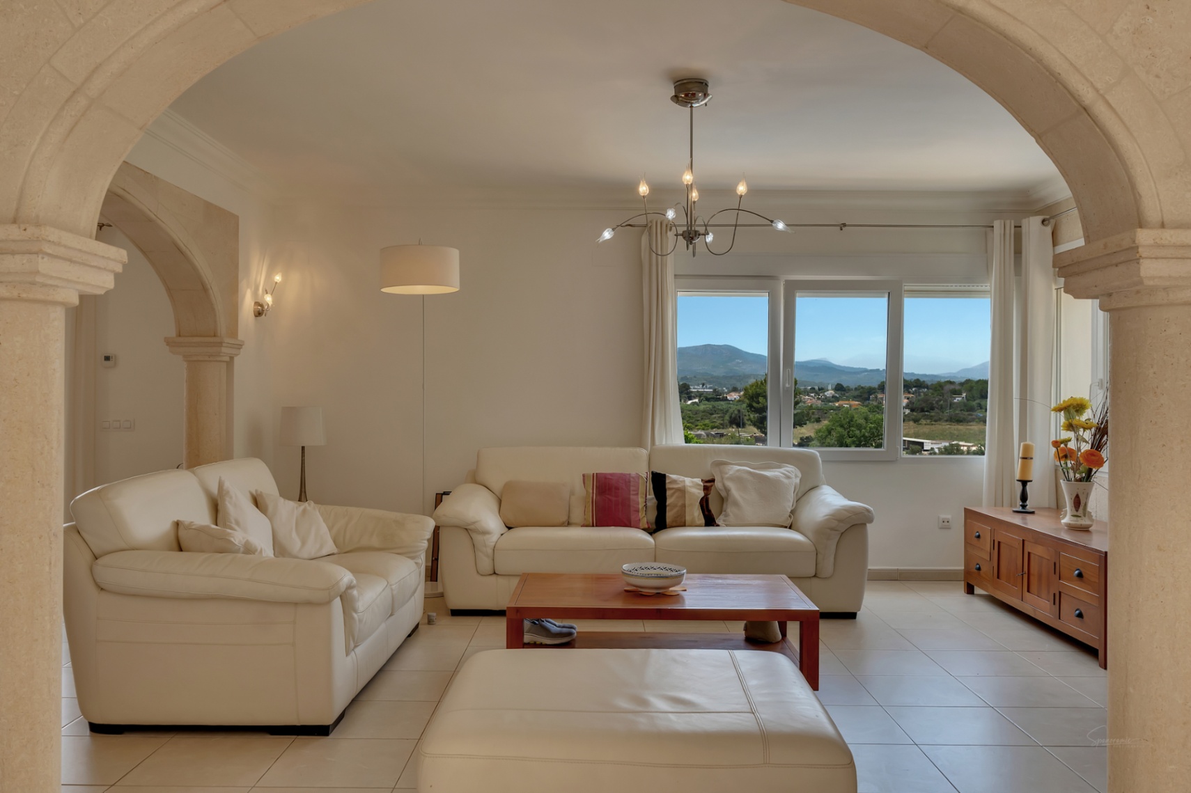 Salón con sofás de cuero blanco enmarcado por arcos de piedra tosca, con vistas panorámicas al valle y las montañas a través de un gran ventanal.