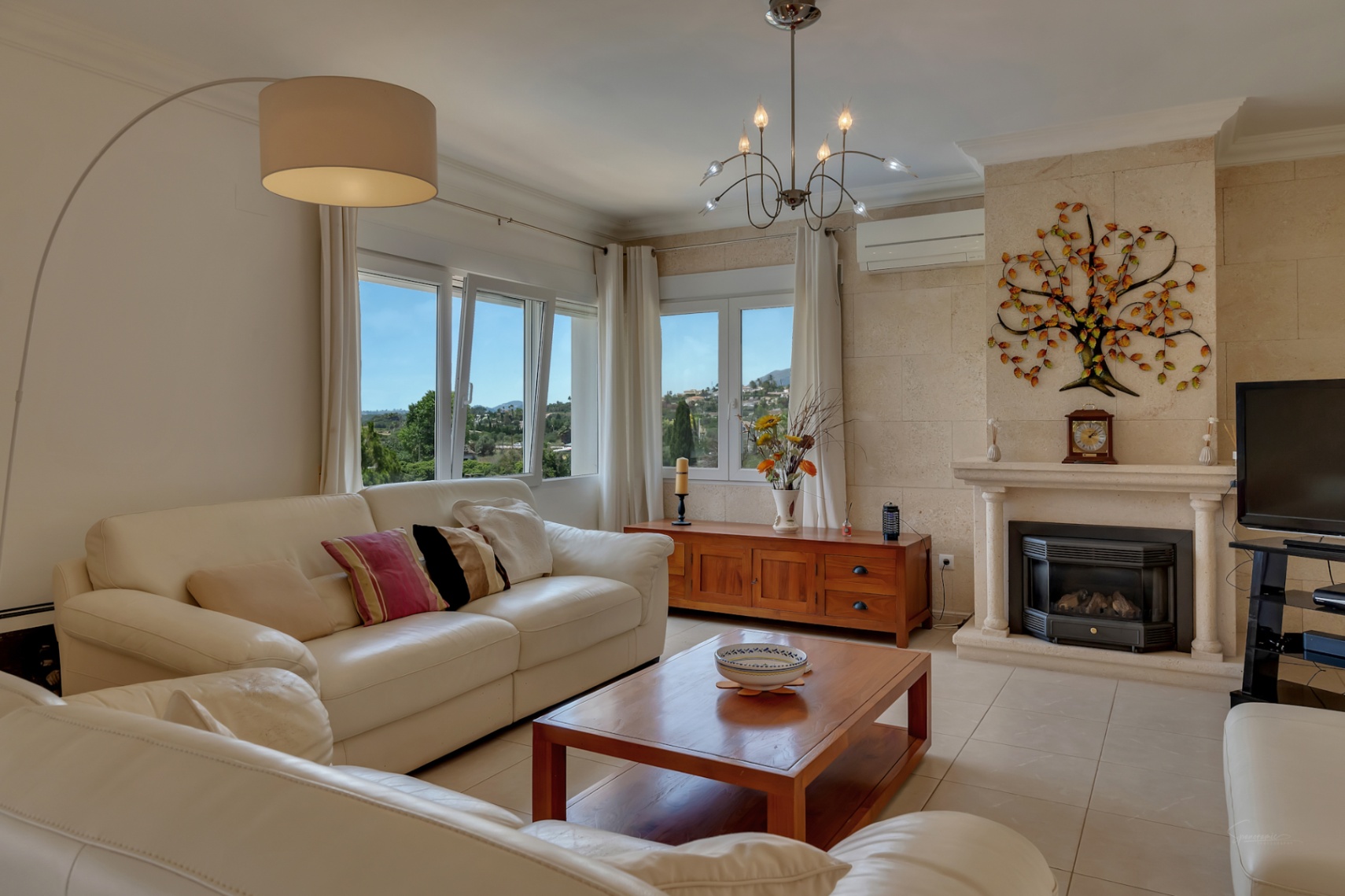 Salón luminoso con sofás de cuero blanco, chimenea de piedra con estufa de leña y amplios ventanales que ofrecen vistas panorámicas al valle de Jávea.