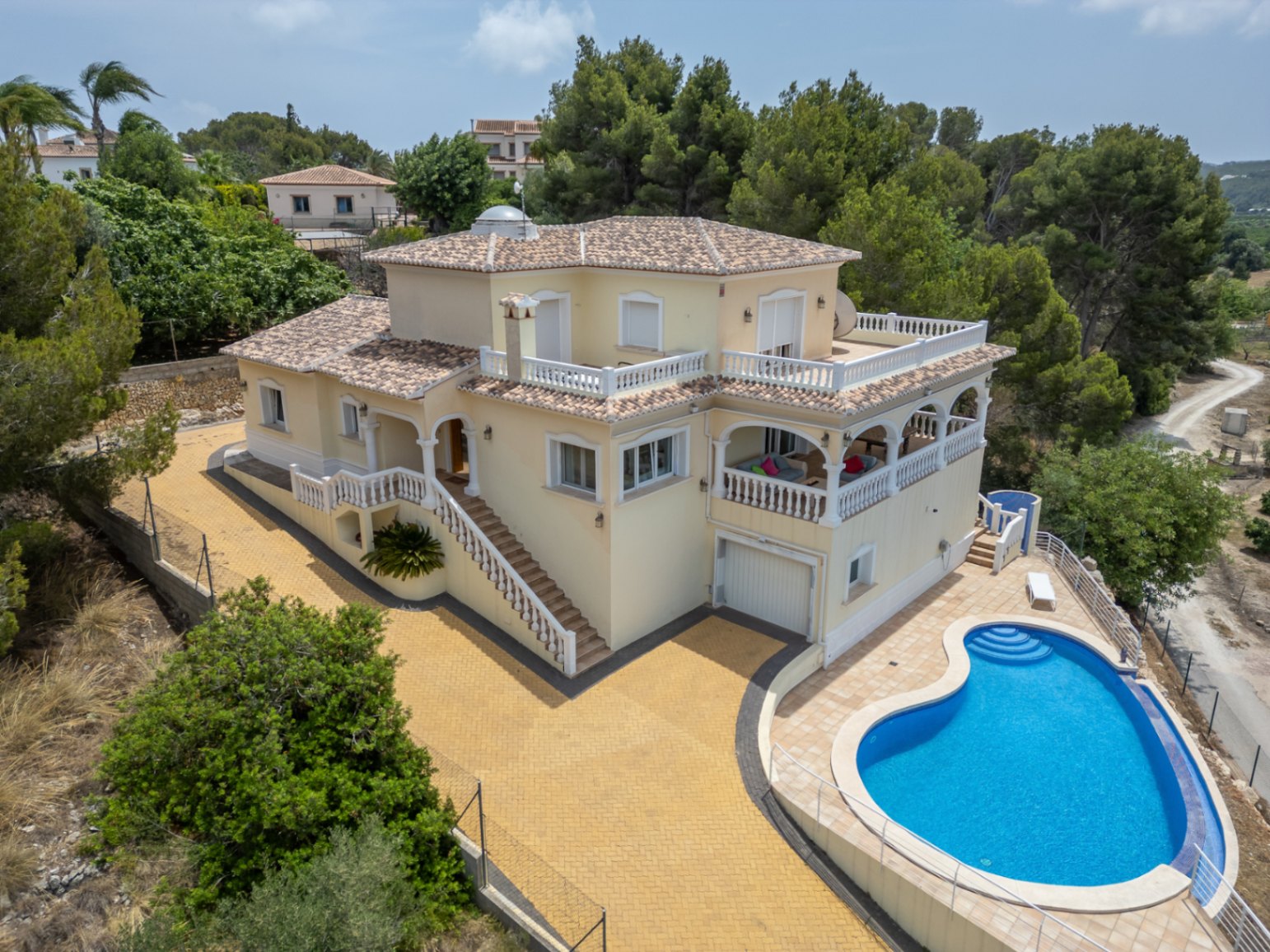 Vista aérea de la villa con fachada crema, naya de arcos y piscina curva, rodeada de amplias terrazas con balaustradas blancas y vegetación mediterránea.