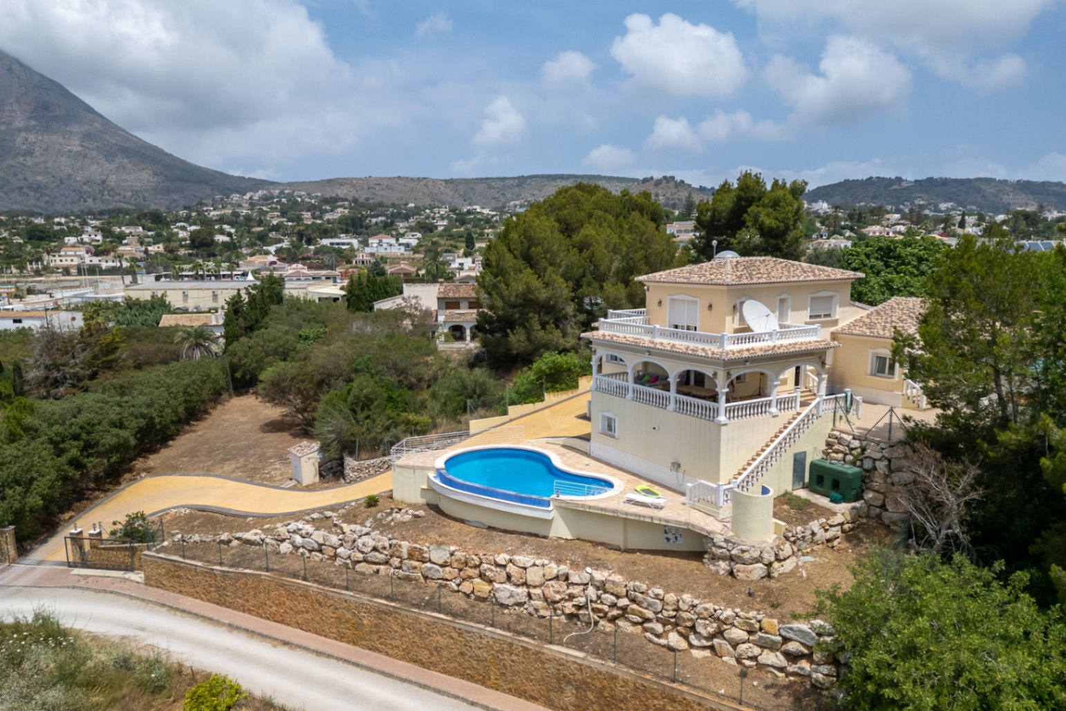 Vista aérea de la villa con piscina curva, amplias terrazas con arcos y balaustradas blancas, y vistas despejadas al Montgó y al valle circundante.
