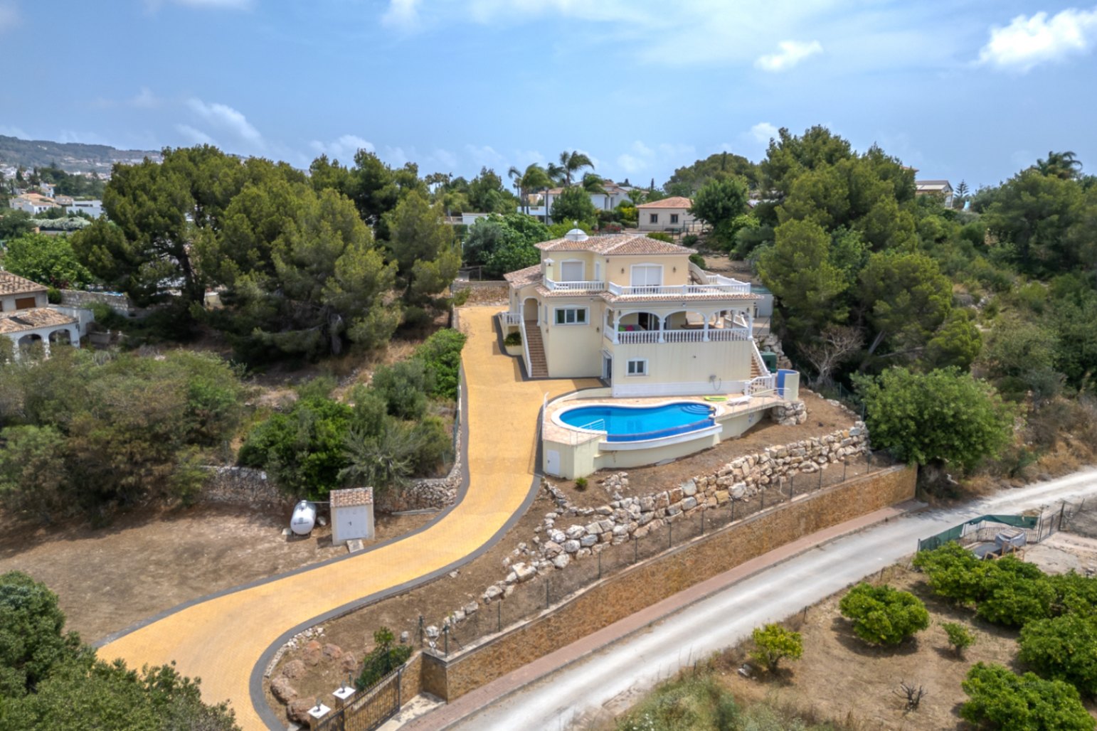 Vista aérea de la villa elevada con piscina infinita de diseño curvo, amplias terrazas y un extenso camino de acceso pavimentado rodeado de pinos.