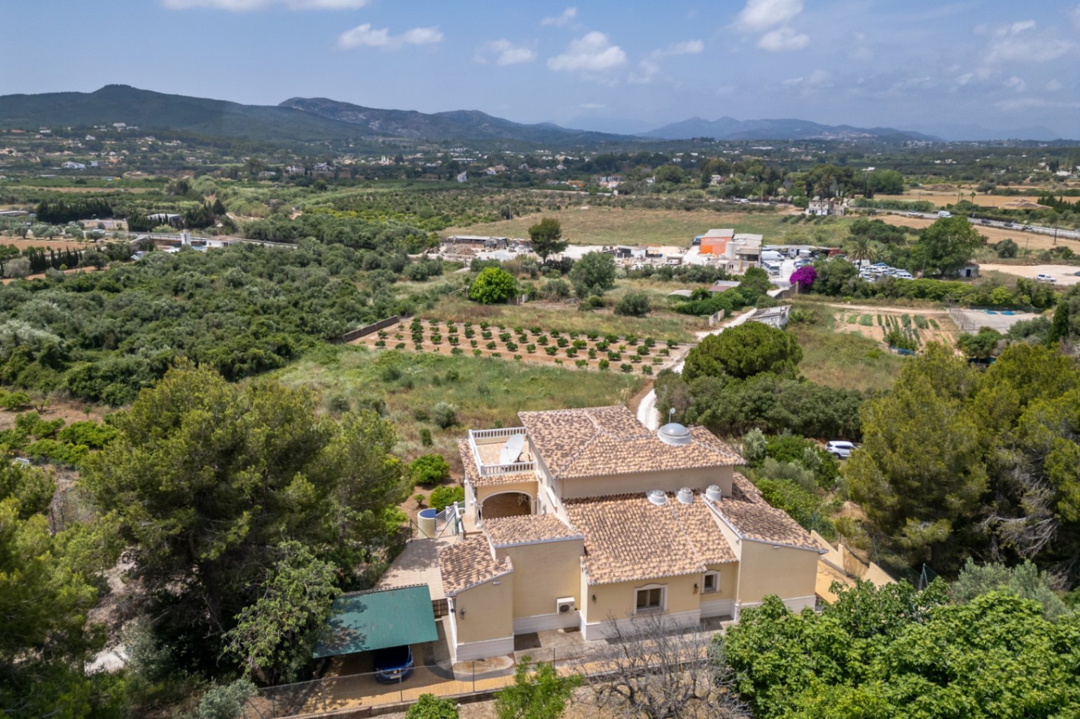 Vista aérea de la villa que muestra su arquitectura con tejados de teja, cúpula central acristalada y claraboyas, rodeada de campos y montañas en Jávea.