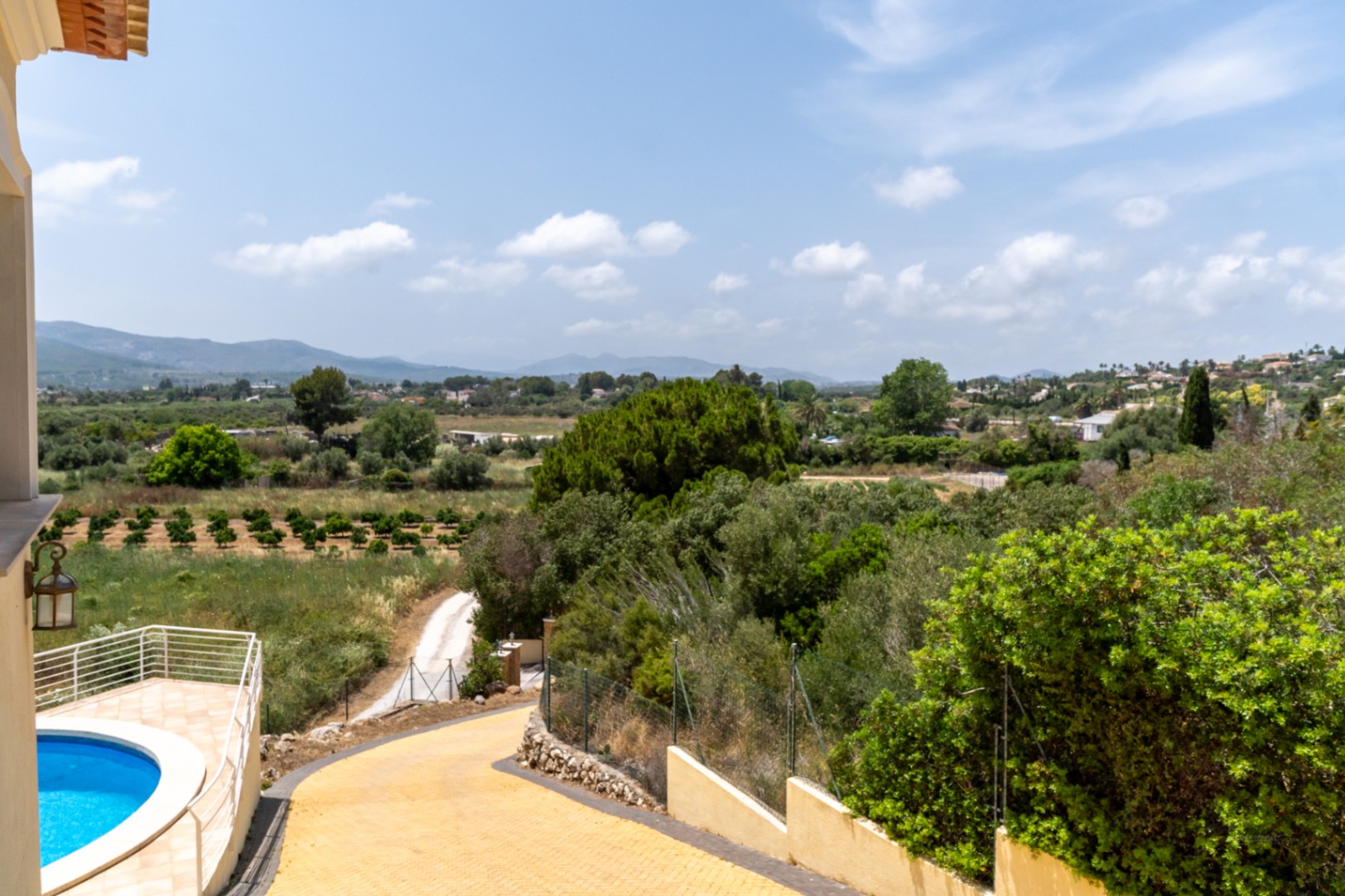 Vista elevada desde la propiedad que muestra el camino de acceso, la piscina circular y una panorámica despejada del valle de Jávea con campos de naranjos.