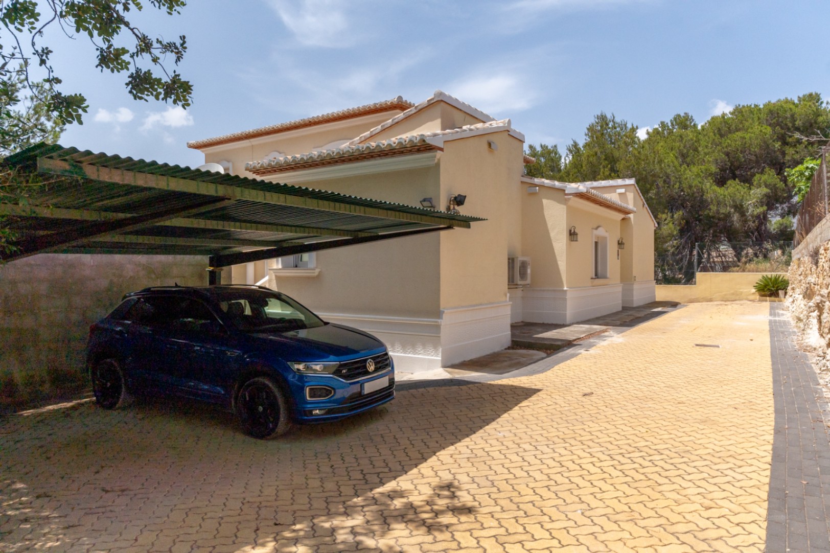 Zona de aparcamiento exterior con pérgola metálica y suelo de adoquines, situada junto a la fachada lateral de la villa con acabados en tonos crema.