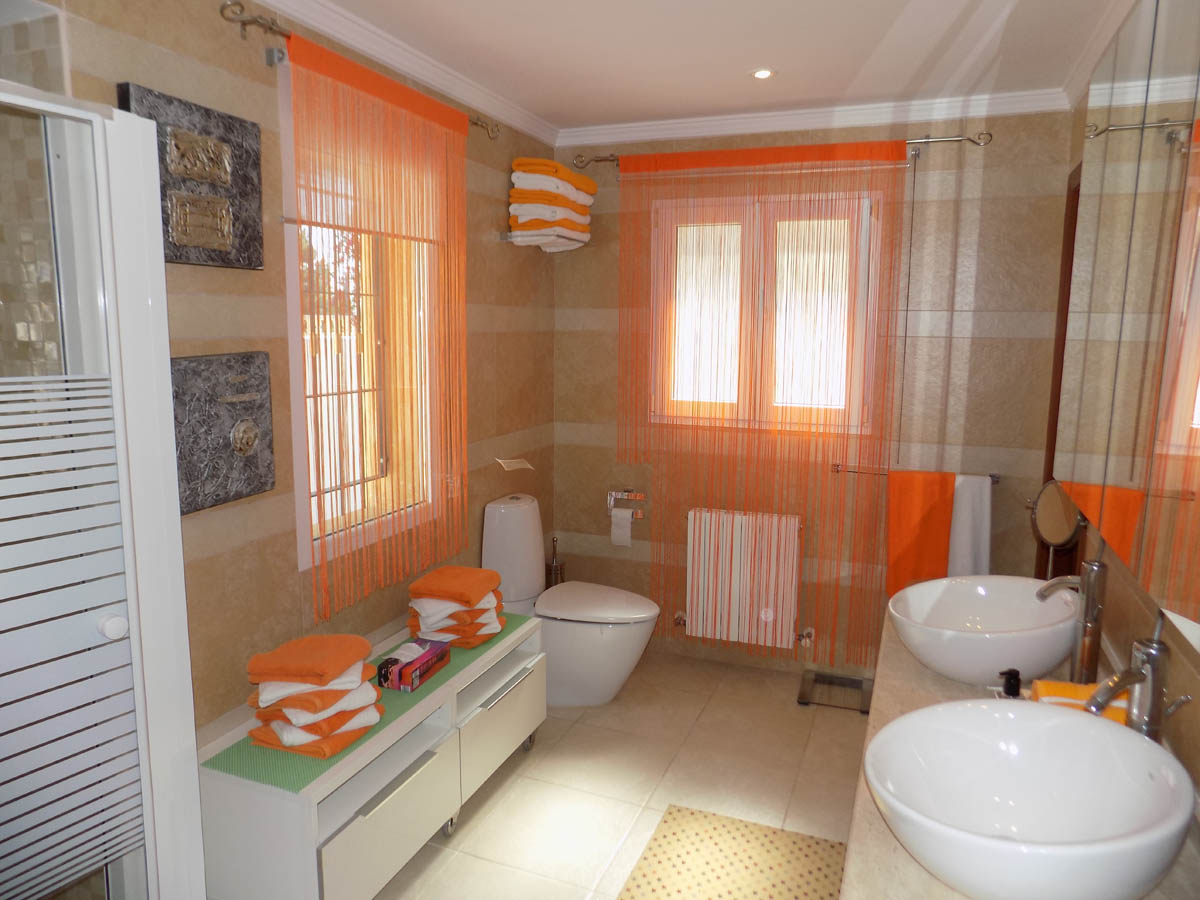 Baño completo con lavabo de doble seno sobre encimera, paredes alicatadas en tonos crema y luz natural a través de amplios ventanales.