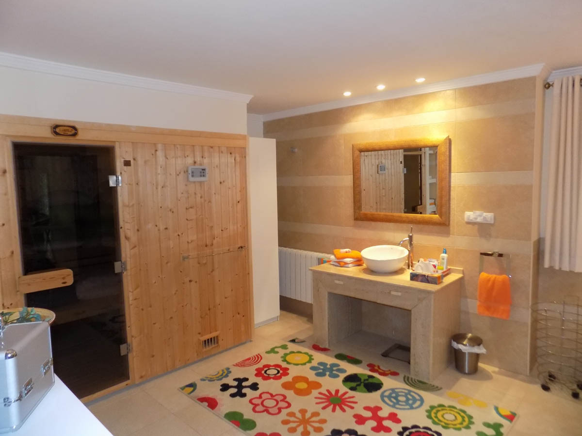 Baño con sauna de madera integrada, lavabo sobre encimera de piedra natural y paredes alicatadas en tonos crema. Incluye iluminación empotrada y radiador.