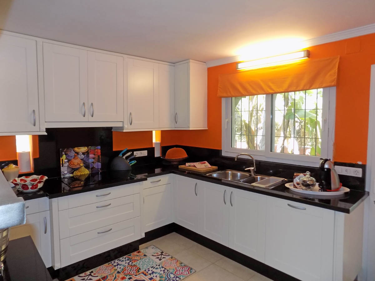 Cocina equipada con armarios blancos, encimera de granito negro y paredes en tono naranja. Incluye fregadero doble bajo ventana con vistas al jardín y suelos cerámicos.
