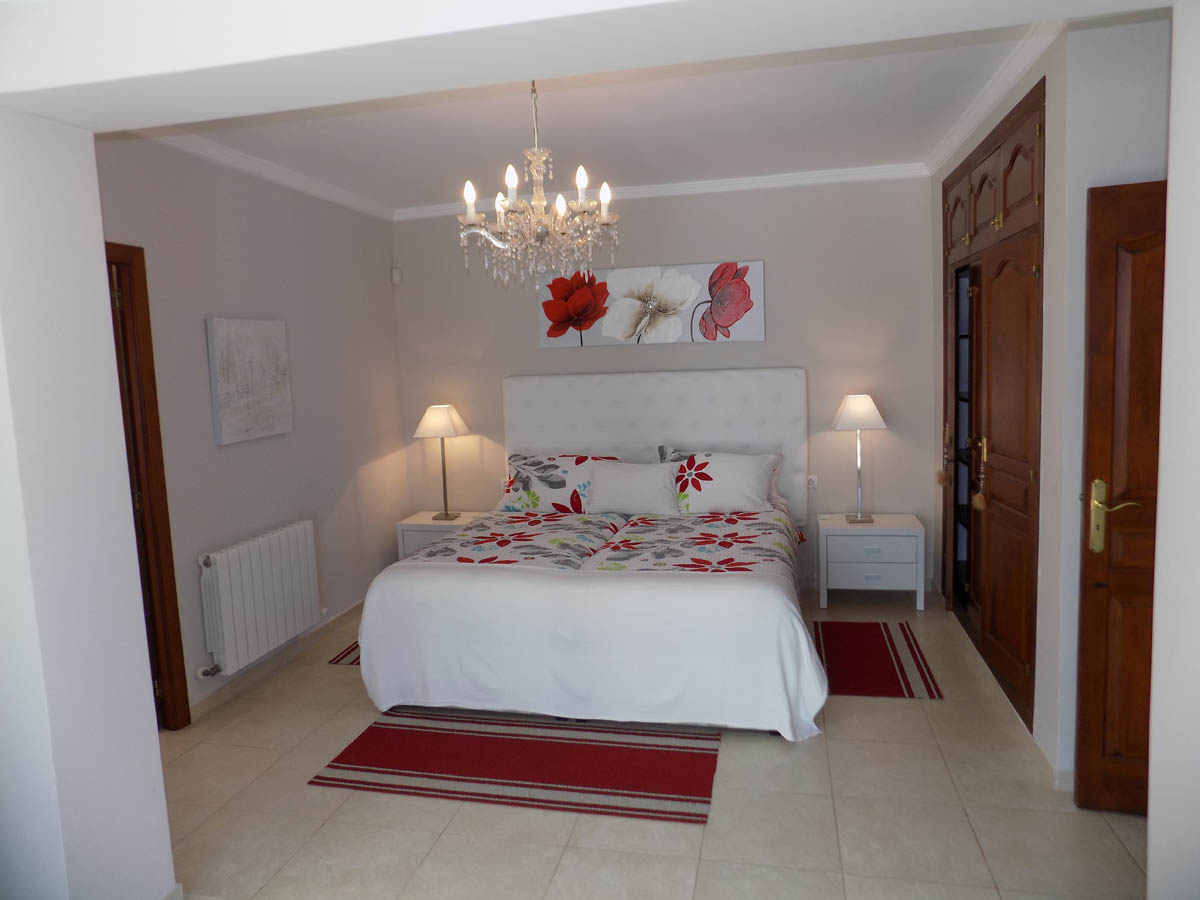Dormitorio principal con armarios empotrados de madera, lámpara de araña clásica, suelos cerámicos y calefacción por radiador en un ambiente luminoso.