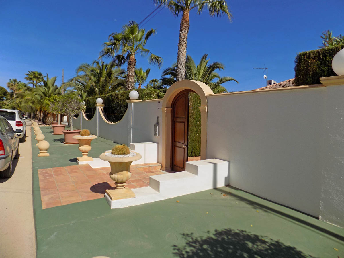 Entrada peatonal con puerta de madera bajo arco de piedra, muro perimetral blanco decorado con maceteros de piedra y cactus, rodeado de vegetación mediterránea.