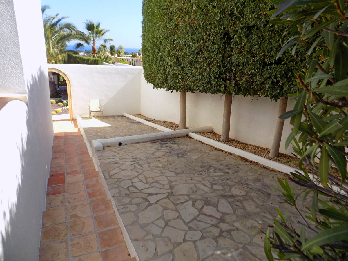Pasillo exterior con pavimento de piedra natural, muros blancos y árboles ornamentales podados que conducen a un arco con vistas al mar y palmeras.