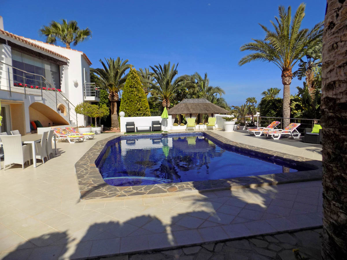 Piscina privada con borde de piedra, amplia terraza de baldosas claras, zona de comedor exterior y un cenador de brezo rodeado de palmeras mediterráneas.