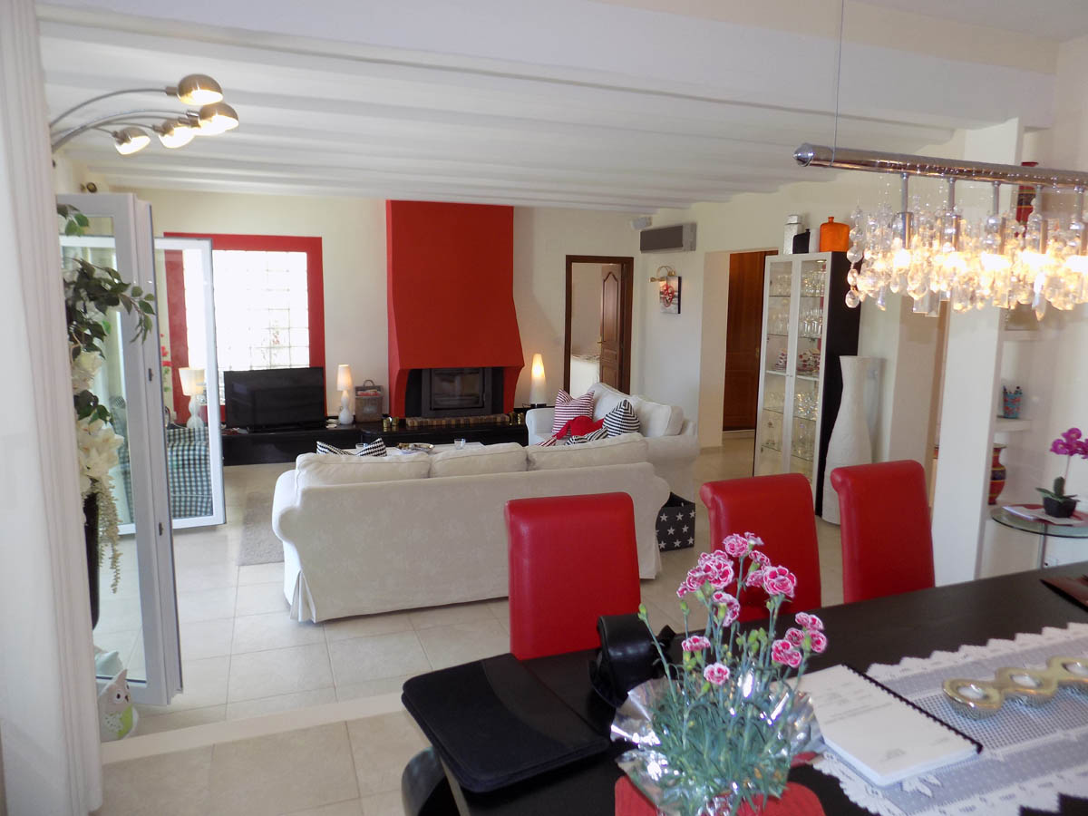 Salón comedor de planta abierta con vigas blancas, chimenea roja distintiva y acceso directo a la terraza acristalada a través de puertas correderas.